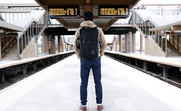 A person carrying a Black 525 Ballistic Western Flyer via the included backpack straps at a rail station platform.